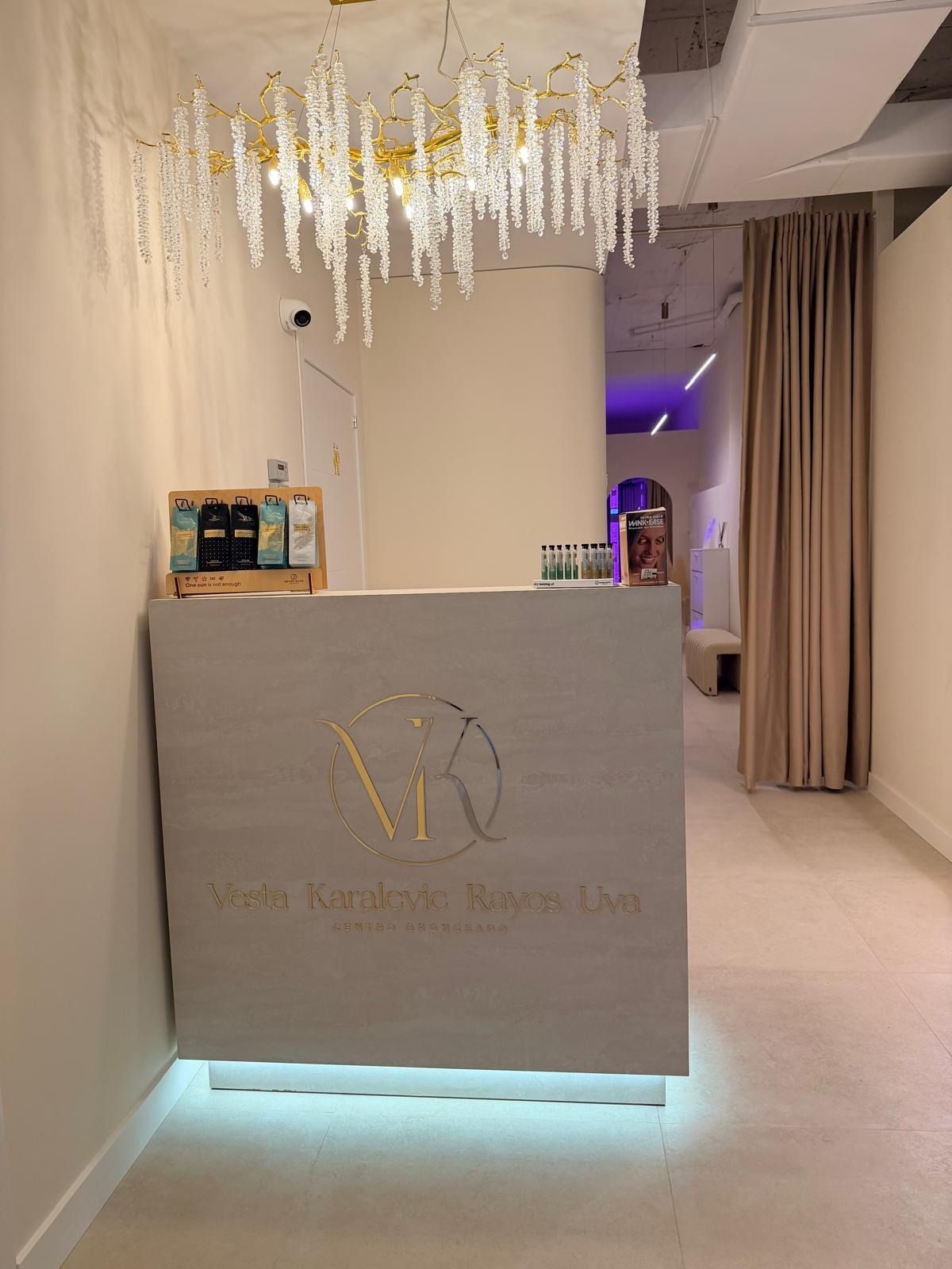 Mostrador de recepción en un spa con una lámpara de araña dorada, texturas de madera clara y cortinas en tonos neutros.