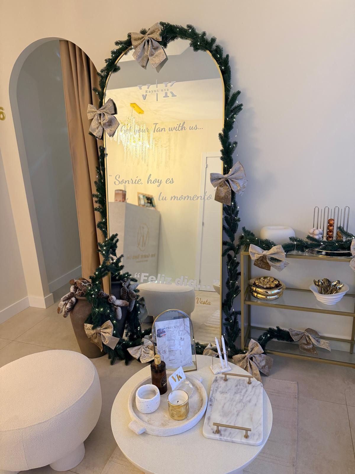 Un espejo arqueado decorado con guirnaldas y lazos navideños, junto a una mesita con velas y una estantería