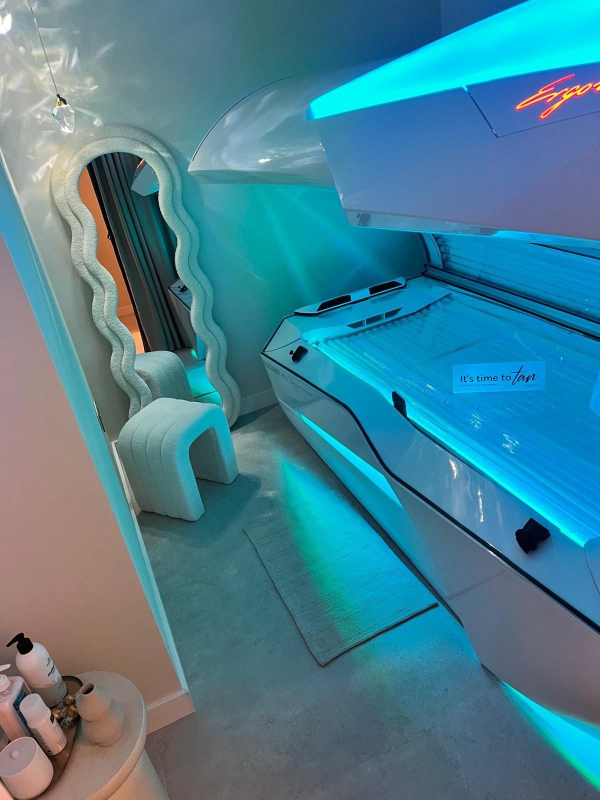 Una cama de bronceado blanca de estilo futurista, iluminada con luces de neón azul, junto a un espejo ondulado