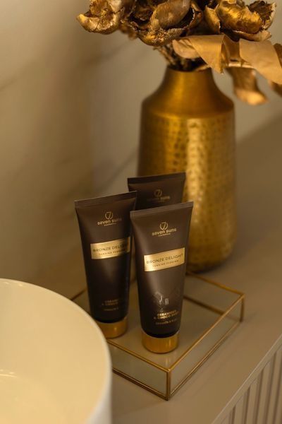 Tres tubos de loción de color marrón oscuro reposan sobre una bandeja dorada junto a un jarrón metálico decorativo