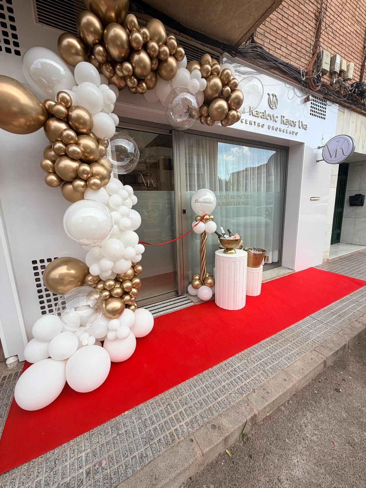 Una alfombra roja conduce a la entrada de una tienda decorada con un gran arco de globos blancos y dorados