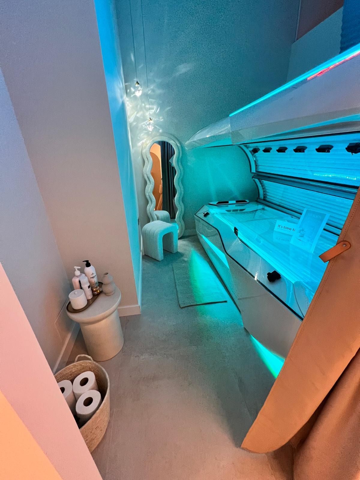 Cama de bronceado en una habitación con iluminación azul, un espejo ondulado y una pequeña mesa auxiliar con productos.