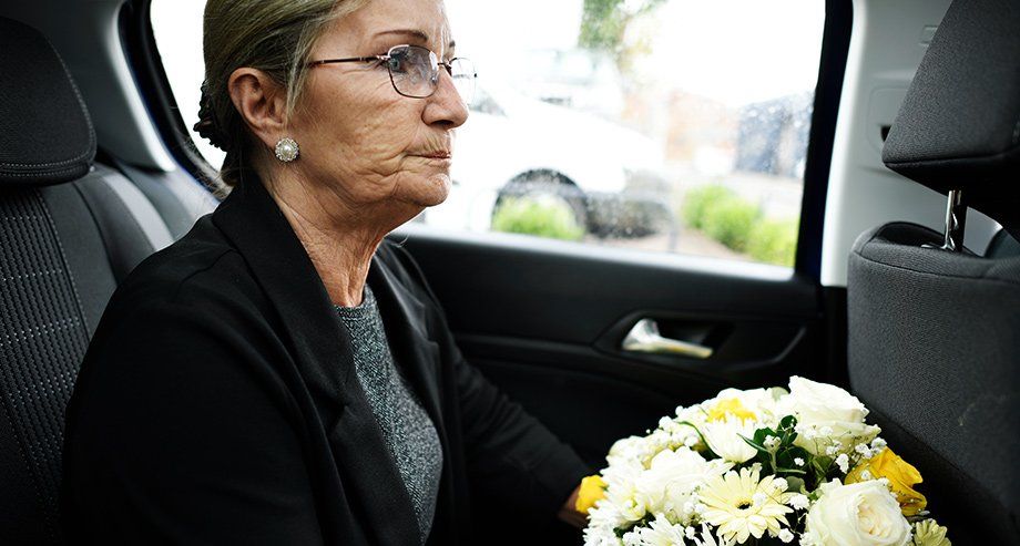 Une femme âgée, portant des habits noirs et tenant un bouquet de fleurs dans les mains, est assise à l'arrière d'une voiture