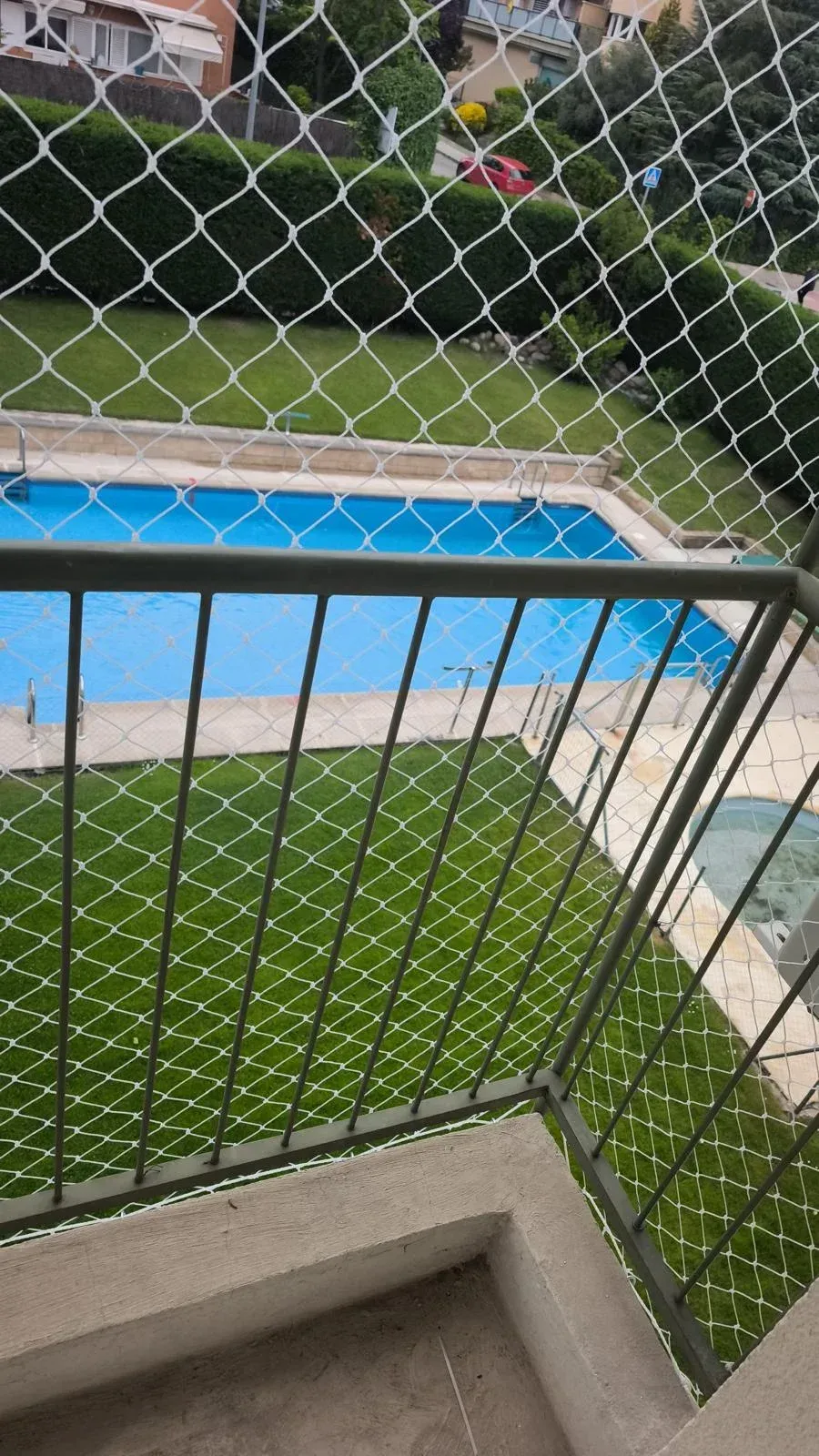 Vista desde un balcón, a través de una valla de tela metálica, de una piscina azul y un patio verde debajo.