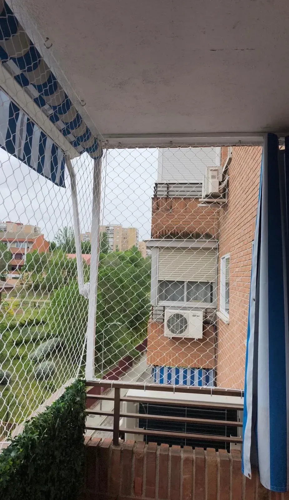 Vista desde el balcón con mosquitera, toldo a rayas, cortina azul y edificio de ladrillo vecino con aire acondicionado.
