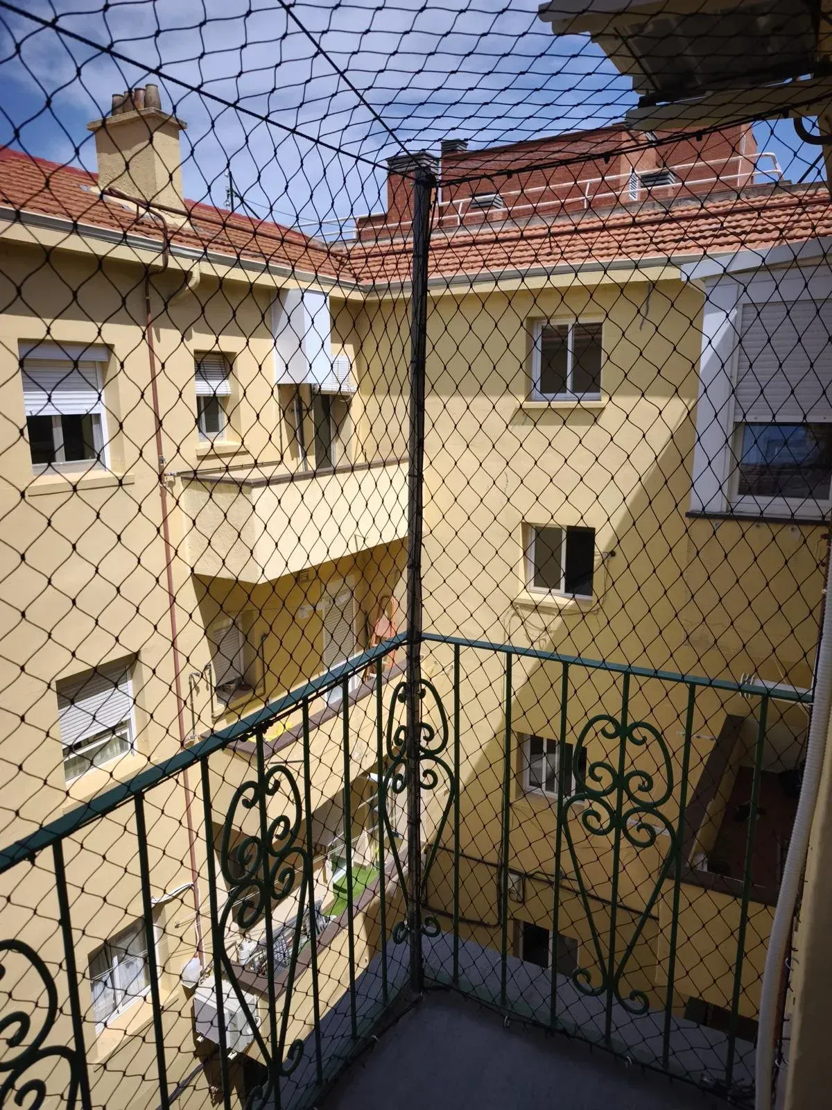Vista desde el balcón de un patio amarillo con techos de tejas rojas, visible a través de una red y una barandilla de hierro forjado.