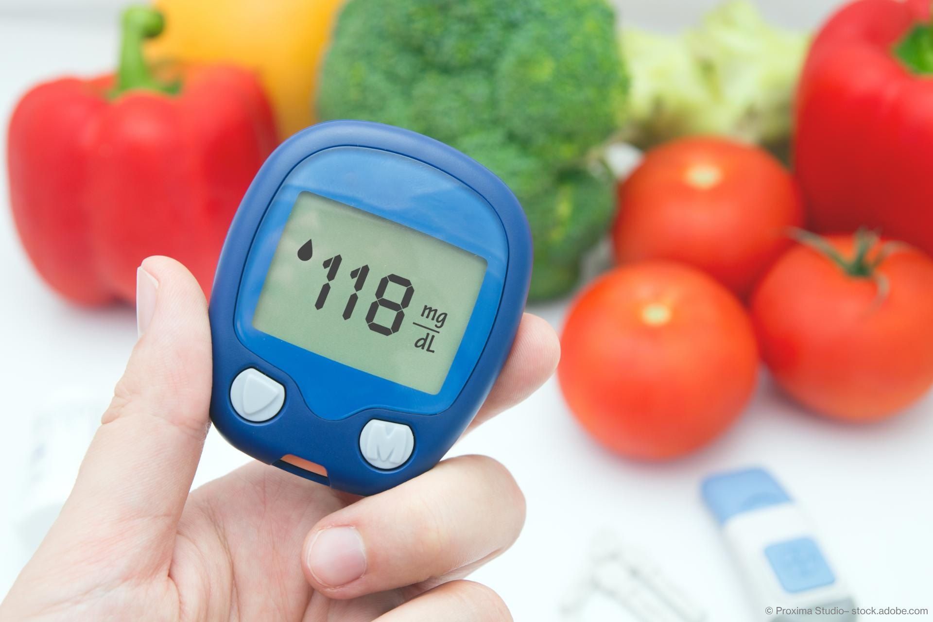 Eine Hand hält ein Blutzuckermessgerät, im Hintergrund liegt ein Diabetes-Heft und Gemüse