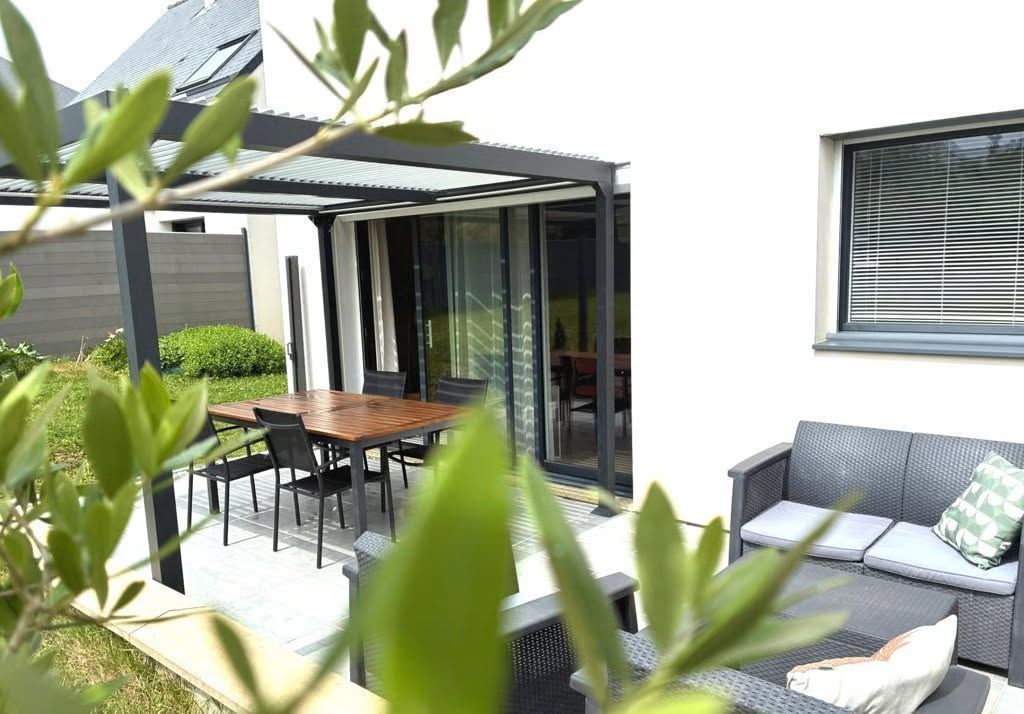 Patio moderne avec table à manger, chaises longues, portes coulissantes en verre et feuillage vert au premier plan