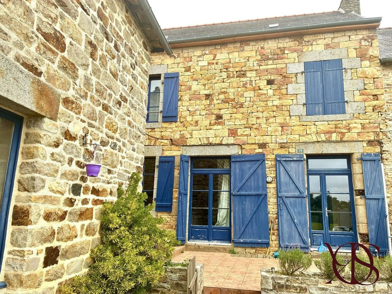 Cour intérieure d'une maison en pierre avec volets bleus, fenêtres et un petit buisson fleuri.