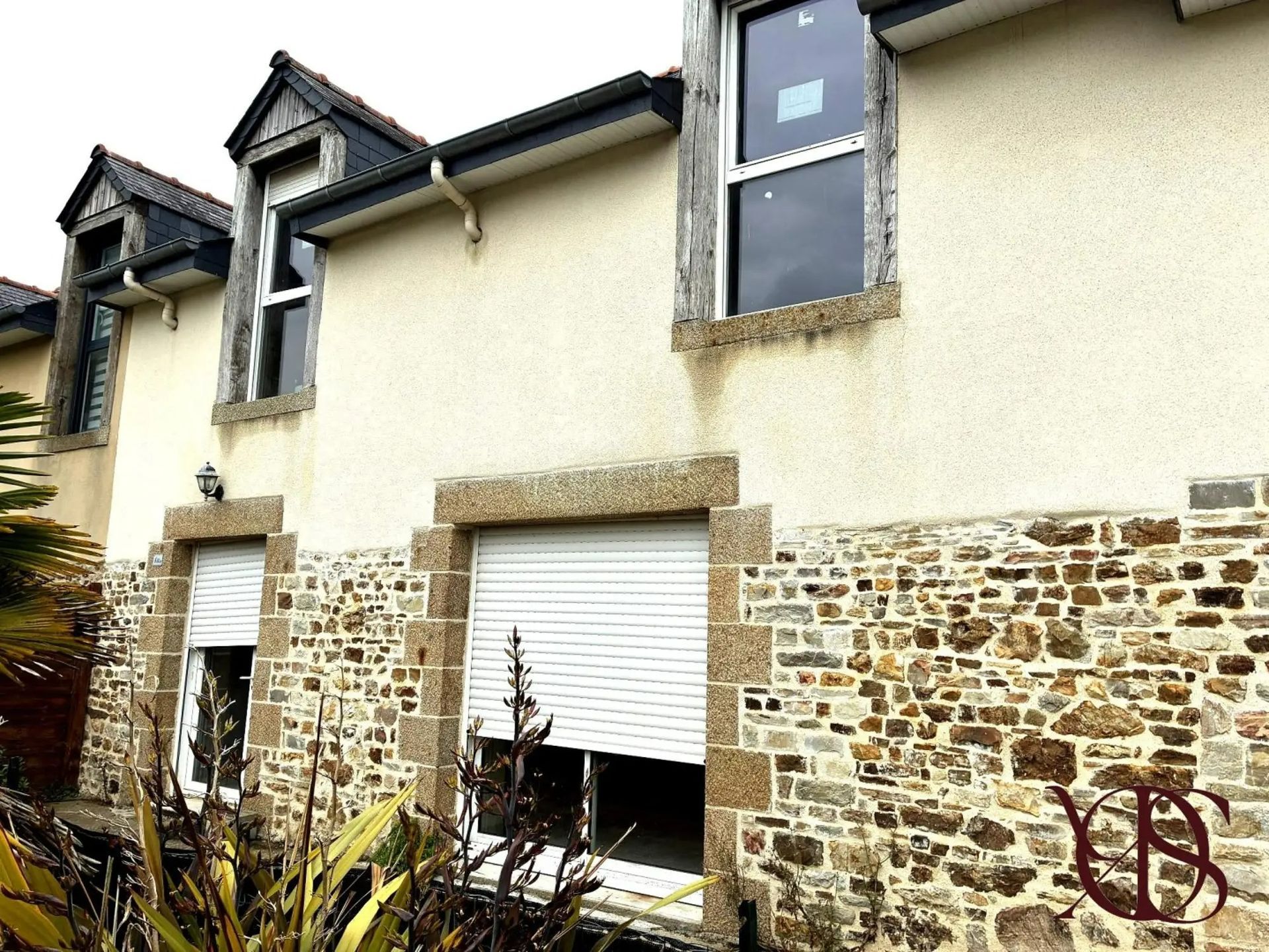 Façade de maison en pierre avec lucarnes et volets fermés, végétation envahissante devant