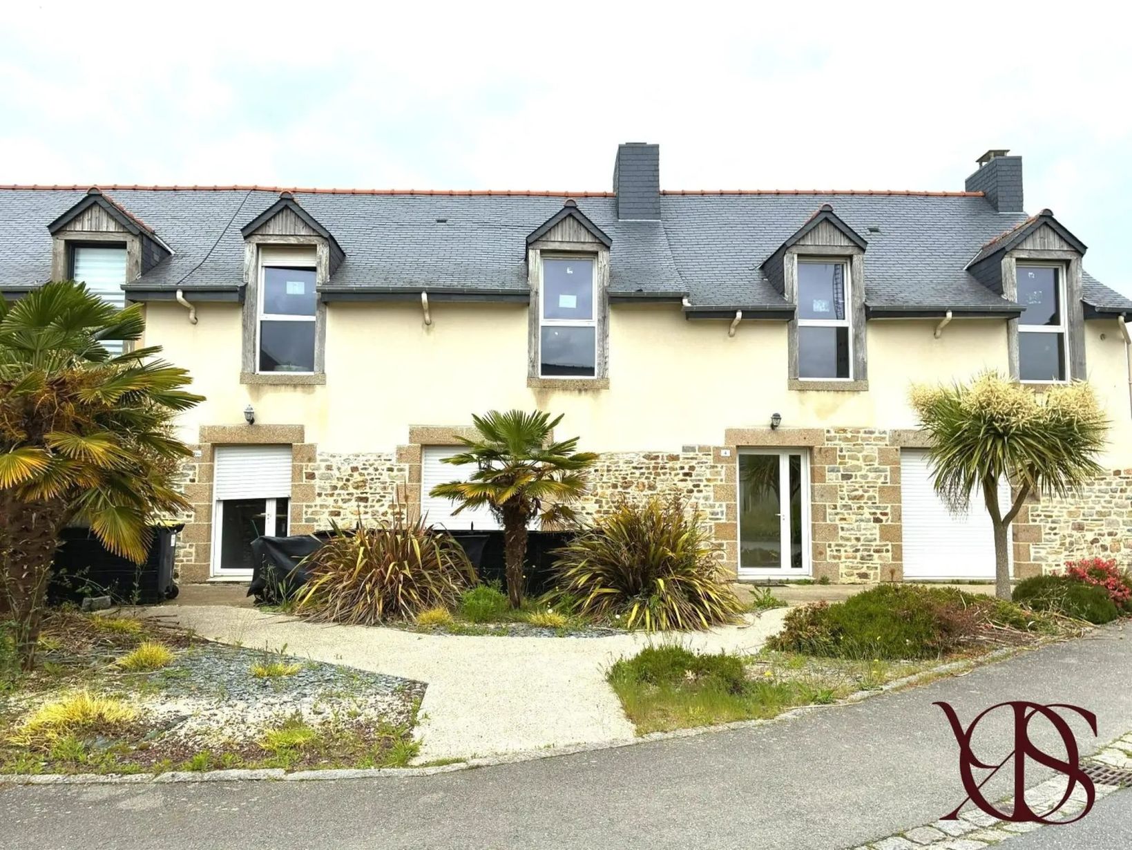 Rangée de maisons de ville couleur crème avec lucarnes à toits sombres, palmiers et allée courbe sous un ciel couvert