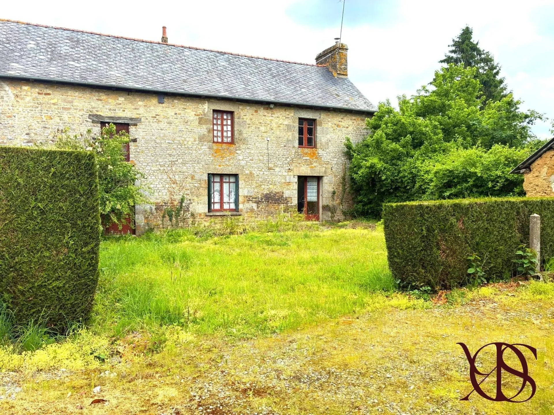 Maisonnette en pierre au toit gris, jardin verdoyant et haies taillées sous un ciel lumineux