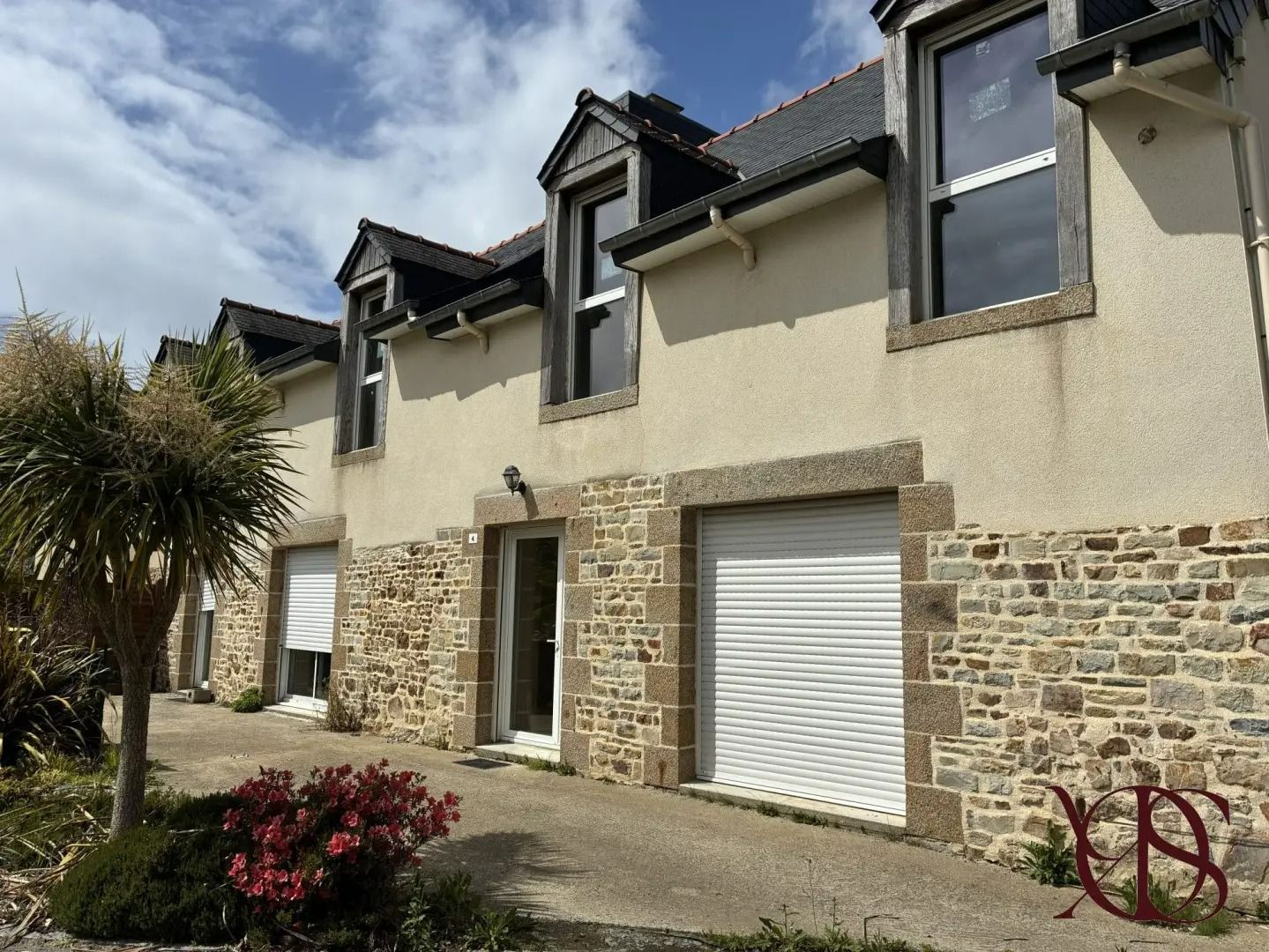 Maison de ville en pierre et en stuc avec portes de garage, palmier et parterre de fleurs sous un ciel nuageux
