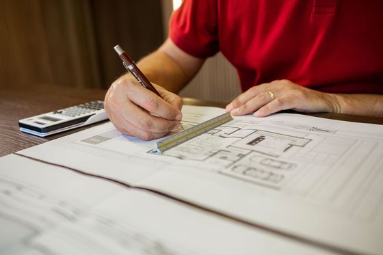 Une personne en chemise rouge dessine les plans d'une maison au crayon à côté d'une calculatrice.