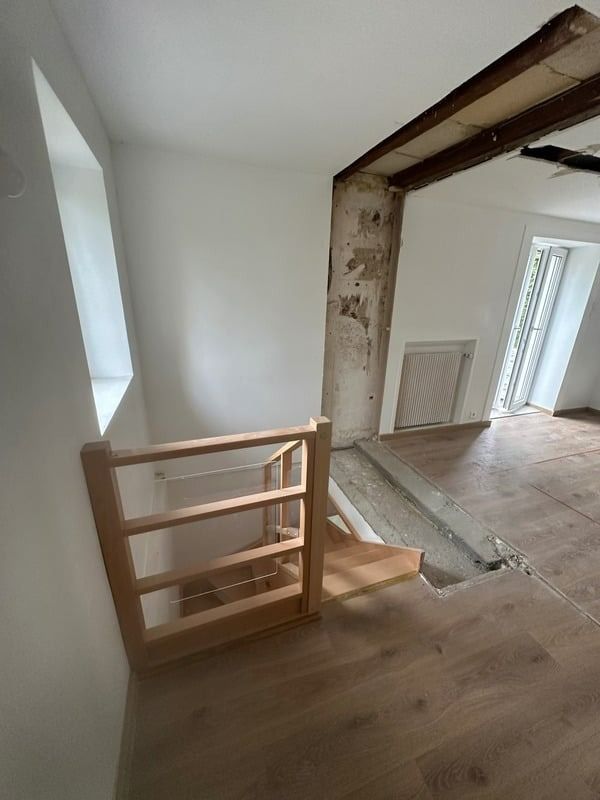 Haut d'un escalier en bois dans un couloir nu avec poutre apparente et porte donnant sur une pièce lumineuse.