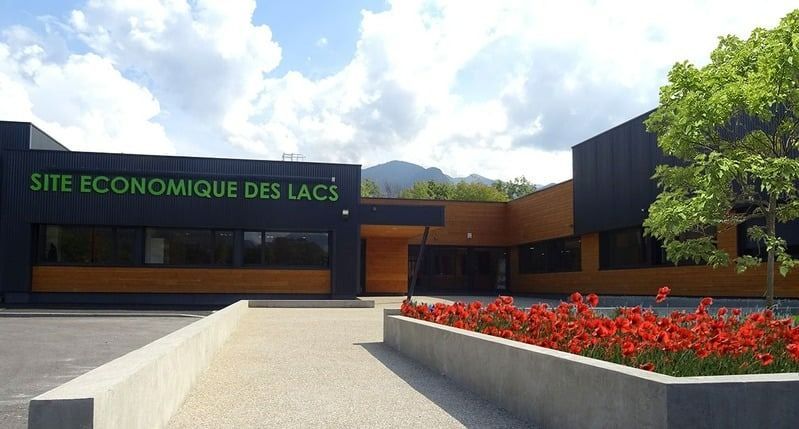 Entrée d'un immeuble moderne avec panneau SITE ÉCONOMIQUE DES LACS et parterres de fleurs rouges.
