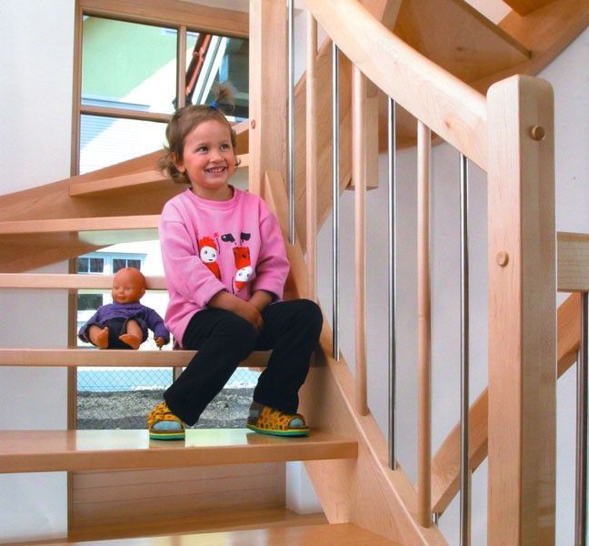 Mädchen sitzt lächelnd mit einer Puppe auf einer Holztreppe. Helles Holzgeländer und Treppe.