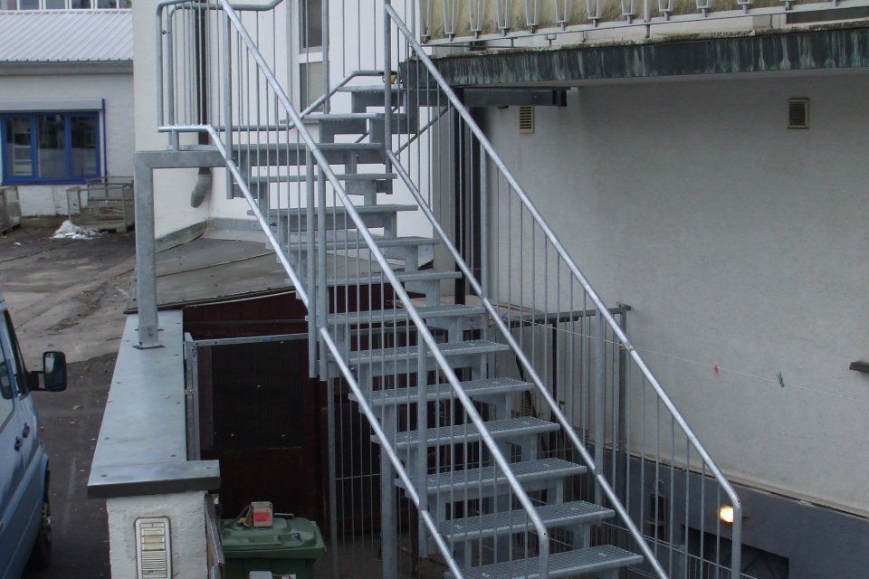 Eine metallene Außentreppe mit Geländer an einem weißen Gebäude führt zu einem Balkon.