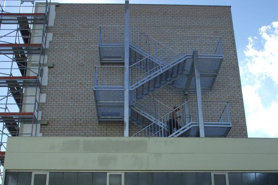Außenansicht eines Gebäudes mit einer metallenen Feuertreppe. Eine Person befindet sich auf der Treppe. An der Seite steht ein Gerüst.