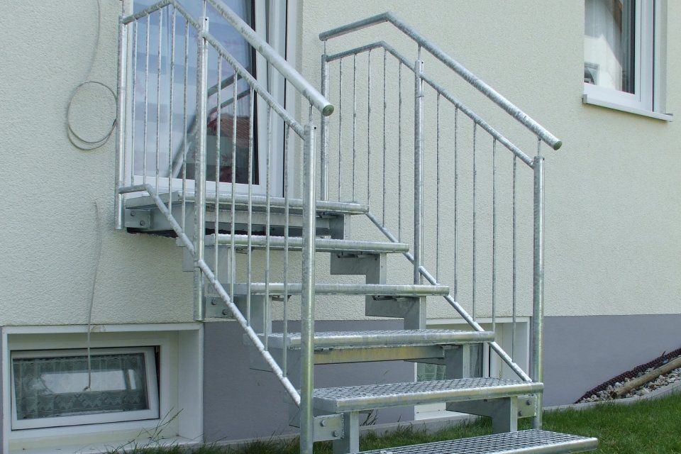 Eine Metalltreppe führt zu einer Tür, die an ein helles Gebäude angebaut ist.