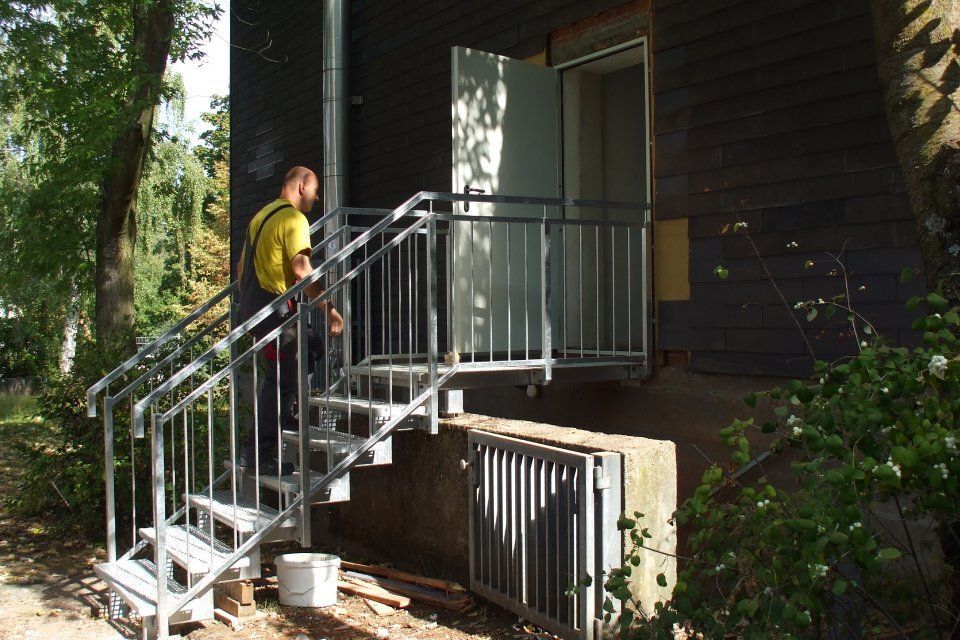 Mann auf einer Metalltreppe, die zu einer Tür führt; dunkle Gebäudefassade.