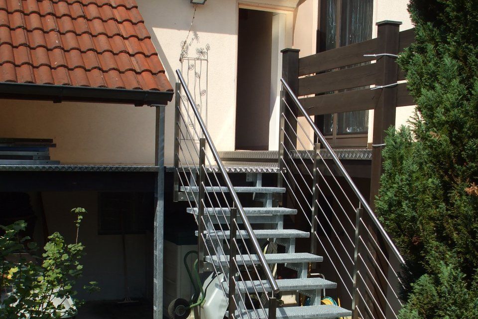 Eine Metalltreppe führt zu einer Tür; an der Wand hängt eine Statue. Braunes Dach, grüne Büsche.