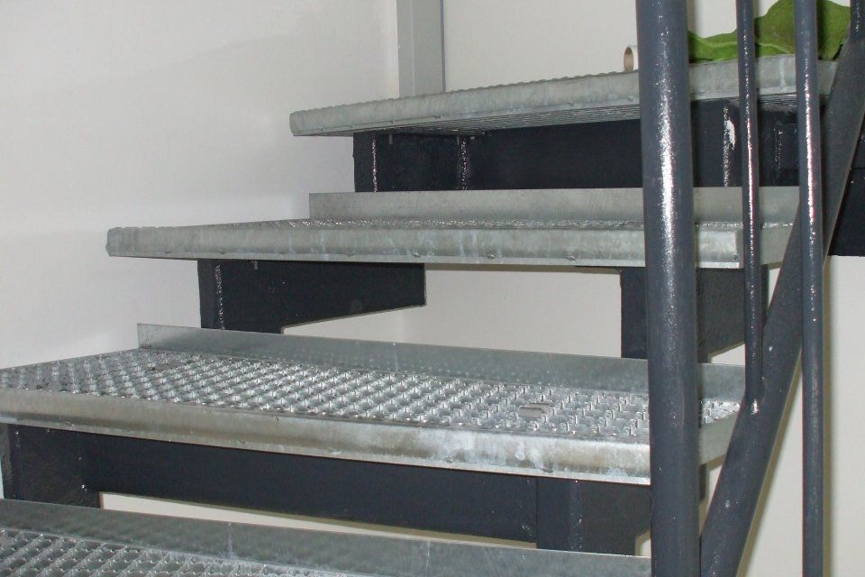 Metalltreppe mit Gitterstufen und dunkelgrauen Stützen vor einer weißen Wand.