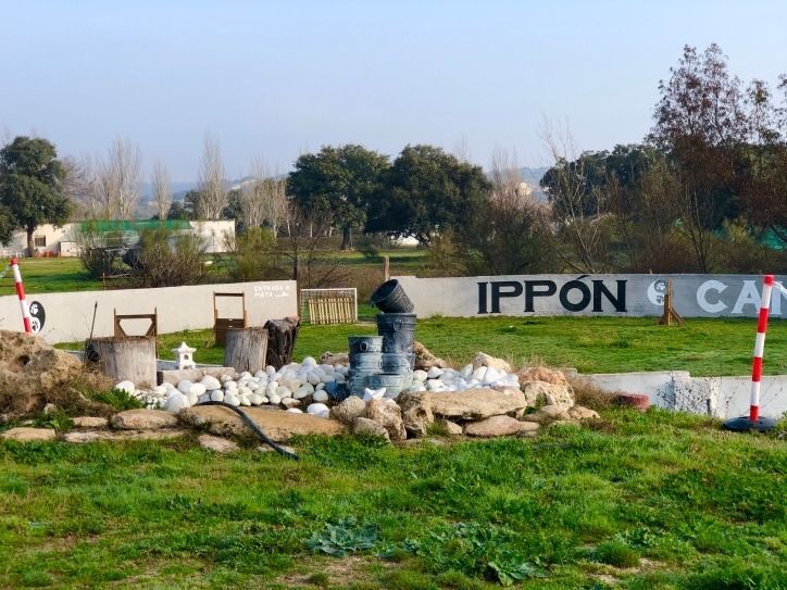 Jardín de rocas con pequeñas estatuas junto a un camino rural, con un letrero de IPPON al fondo.