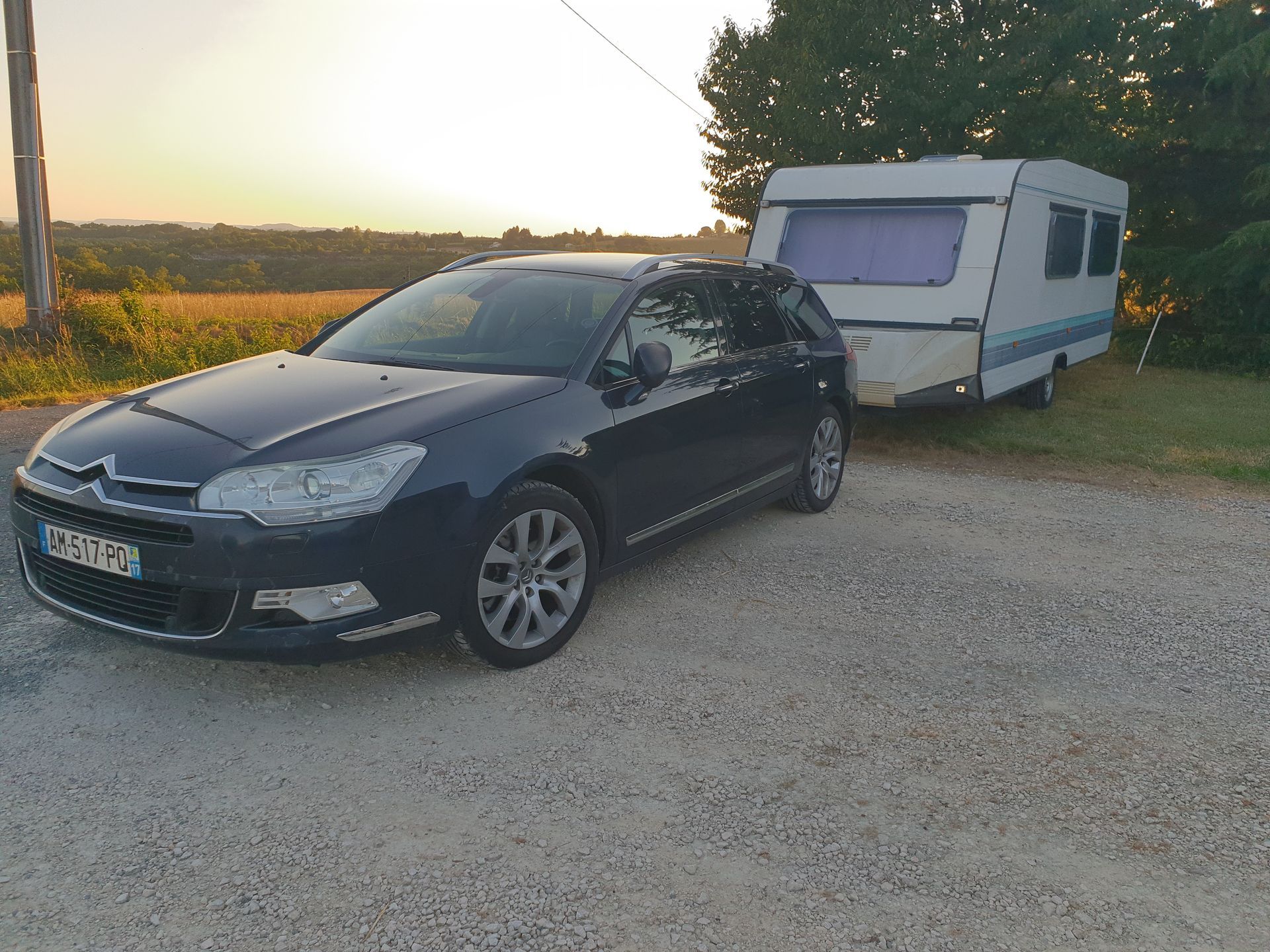 Une Citroën C5 Tourer est garée sur du gravier à côté d'une caravane blanche au coucher du soleil.