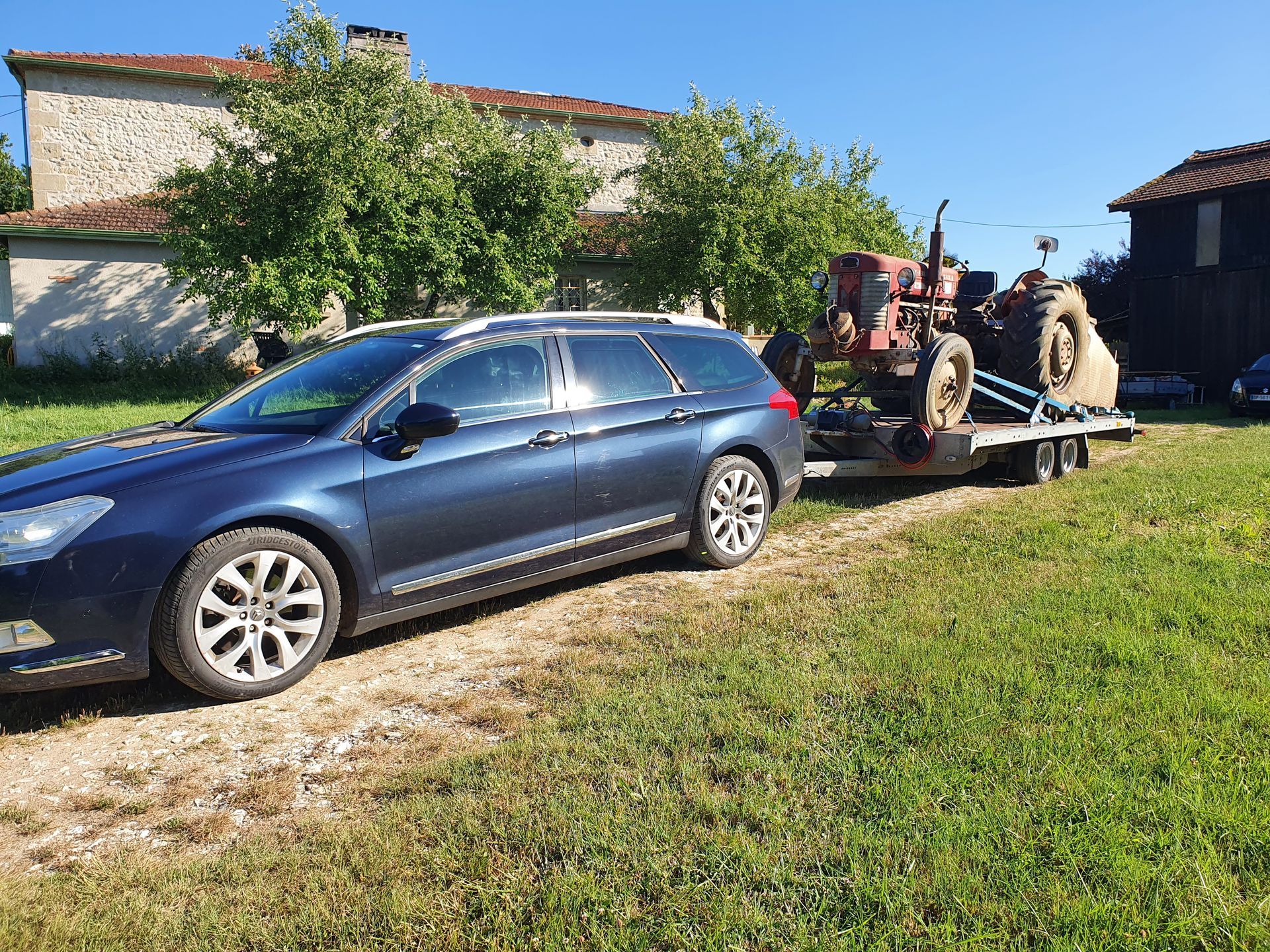 Un break bleu tractant une remorque avec un tracteur dans une cour de campagne ensoleillée.