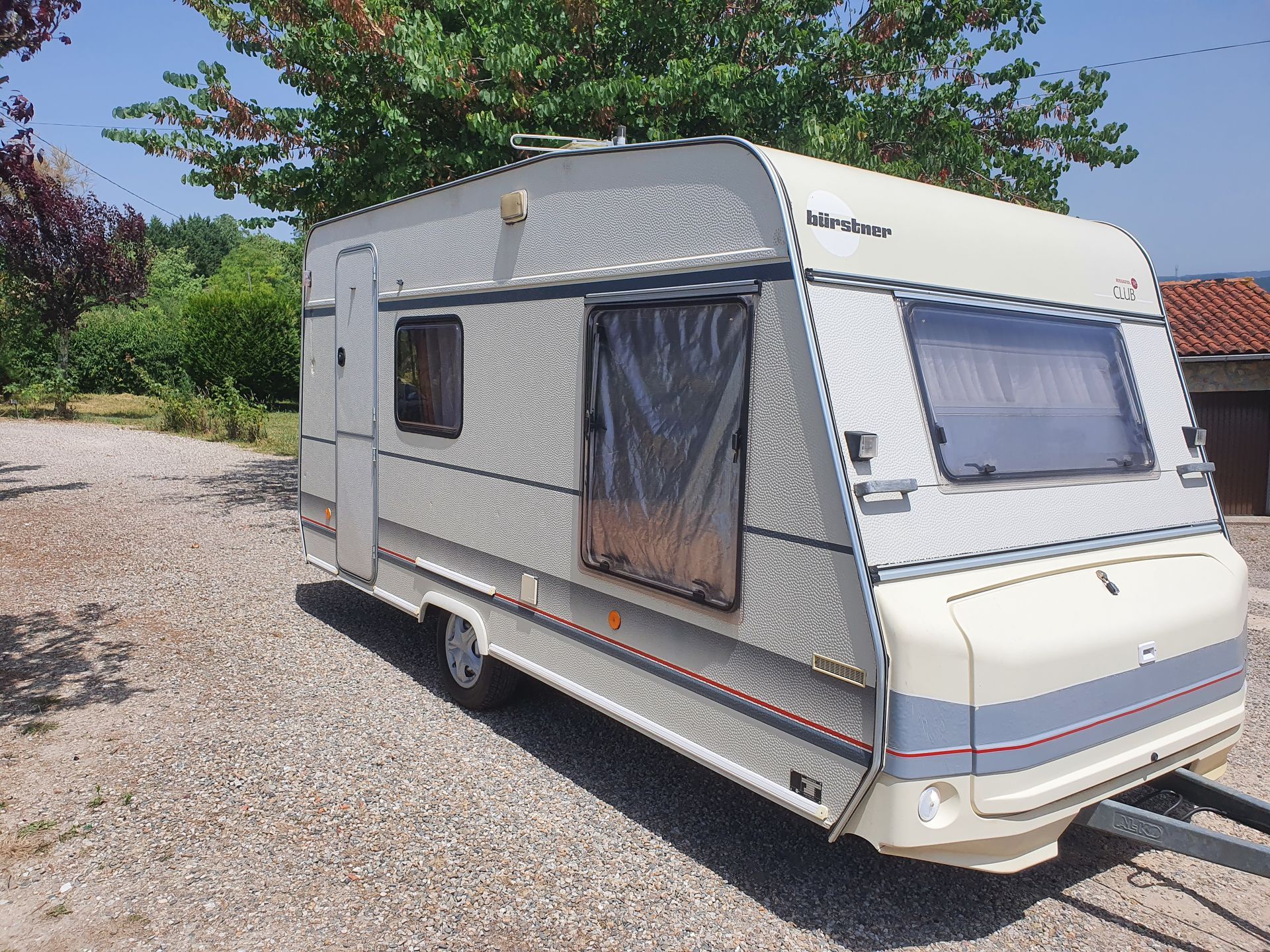 Caravane blanche avec fenêtres latérales est garée sur une allée de gravier à côté d'arbres et de verdure.