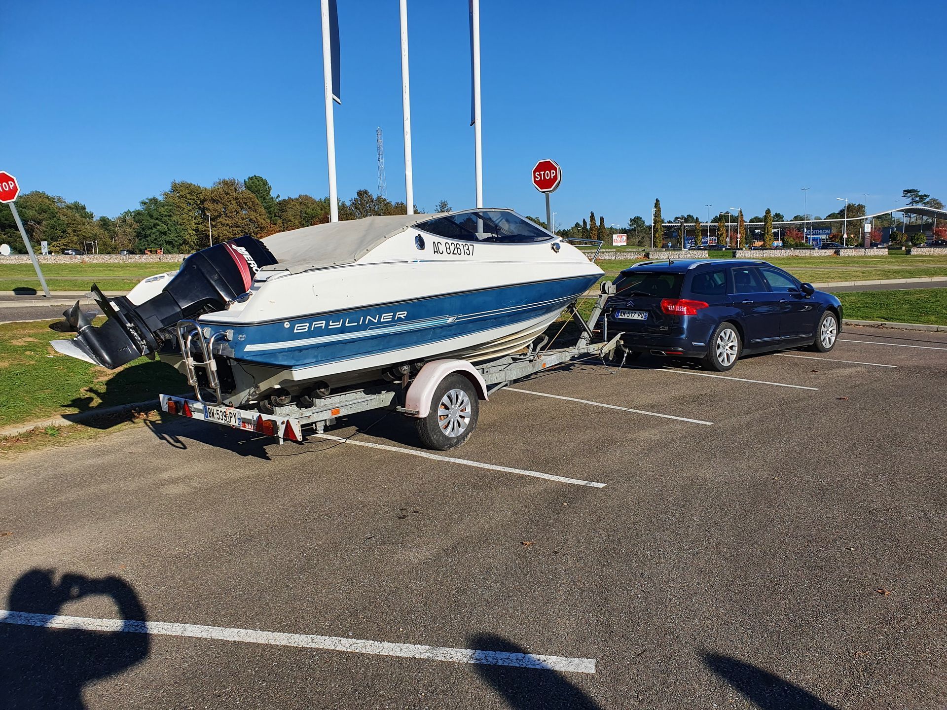 Un bateau sur une remorque adaptée est tracté par une voiture bleue sur un parking par une journée ensoleillée.