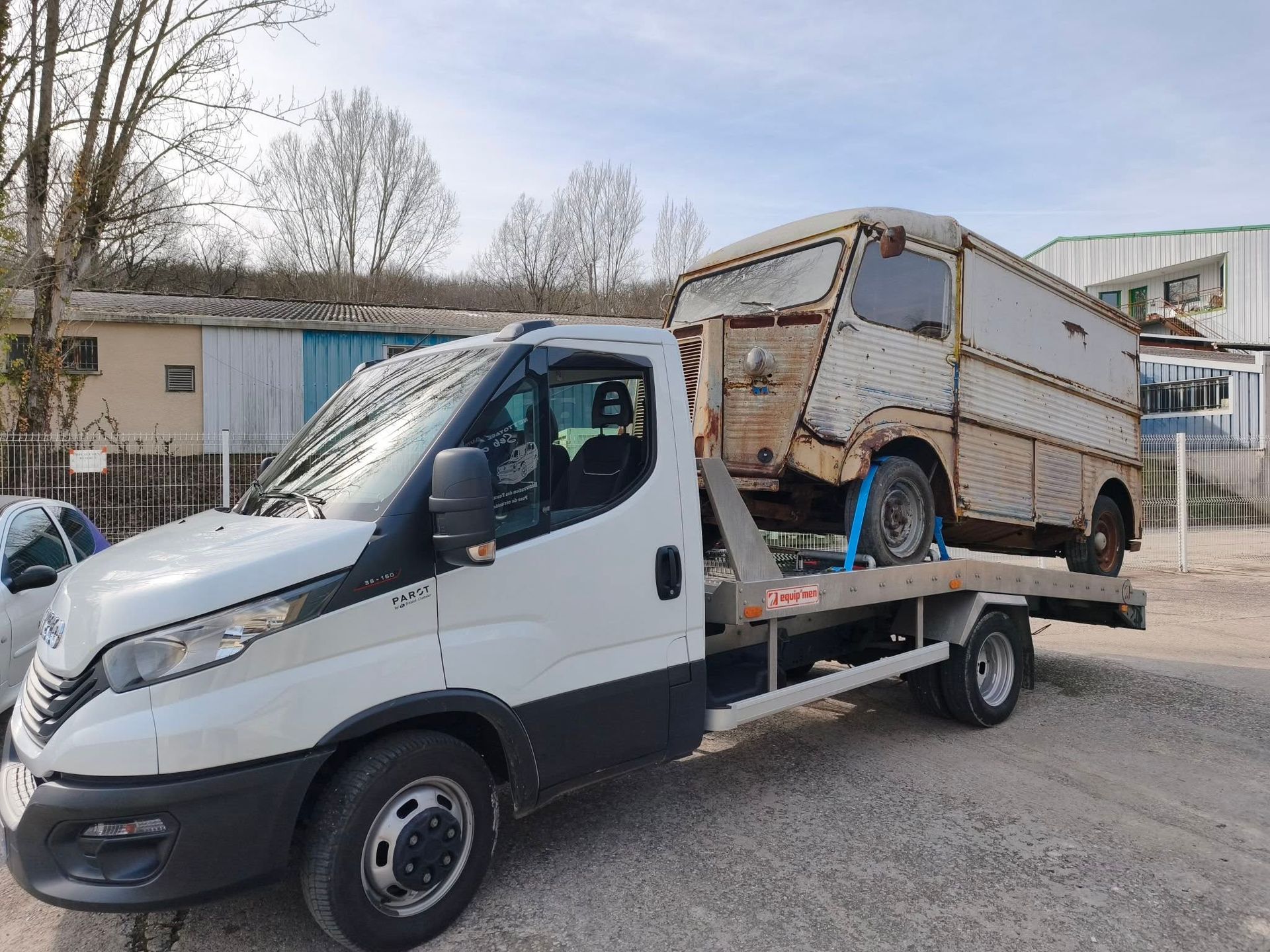 Un camion de dépannage blanc à plateau transporte une vieille camionnette beige dans une allée.
