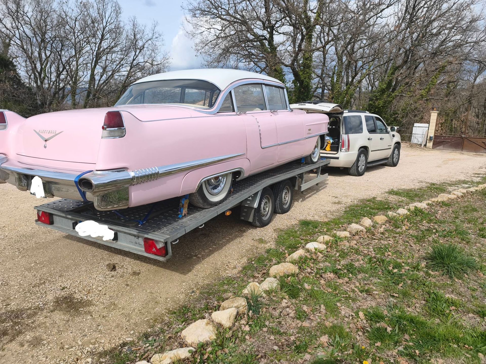 Une Cadillac Fleetwood rose, transportée sur une remorque à plateau par un camion blanc, circule sur un chemin de gravier