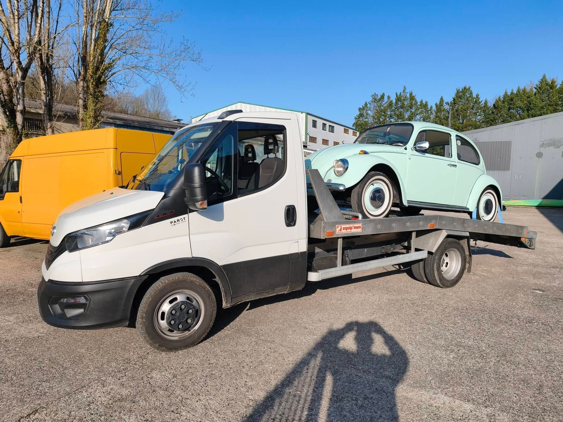 Un camion de dépannage blanc à plateau transporte une voiture ancienne vert menthe sur un parking extérieur ensoleillé.