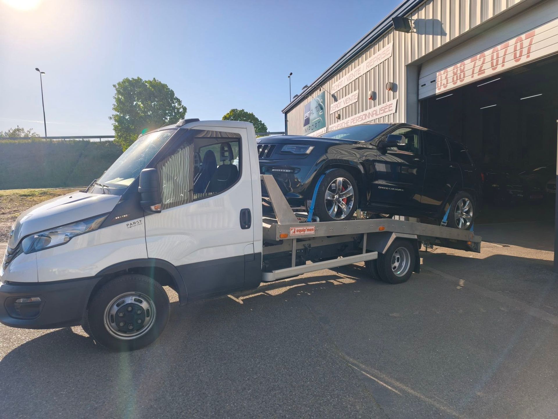 Une dépanneuse blanche à plateau transporte une voiture noire devant un garage en plein soleil.