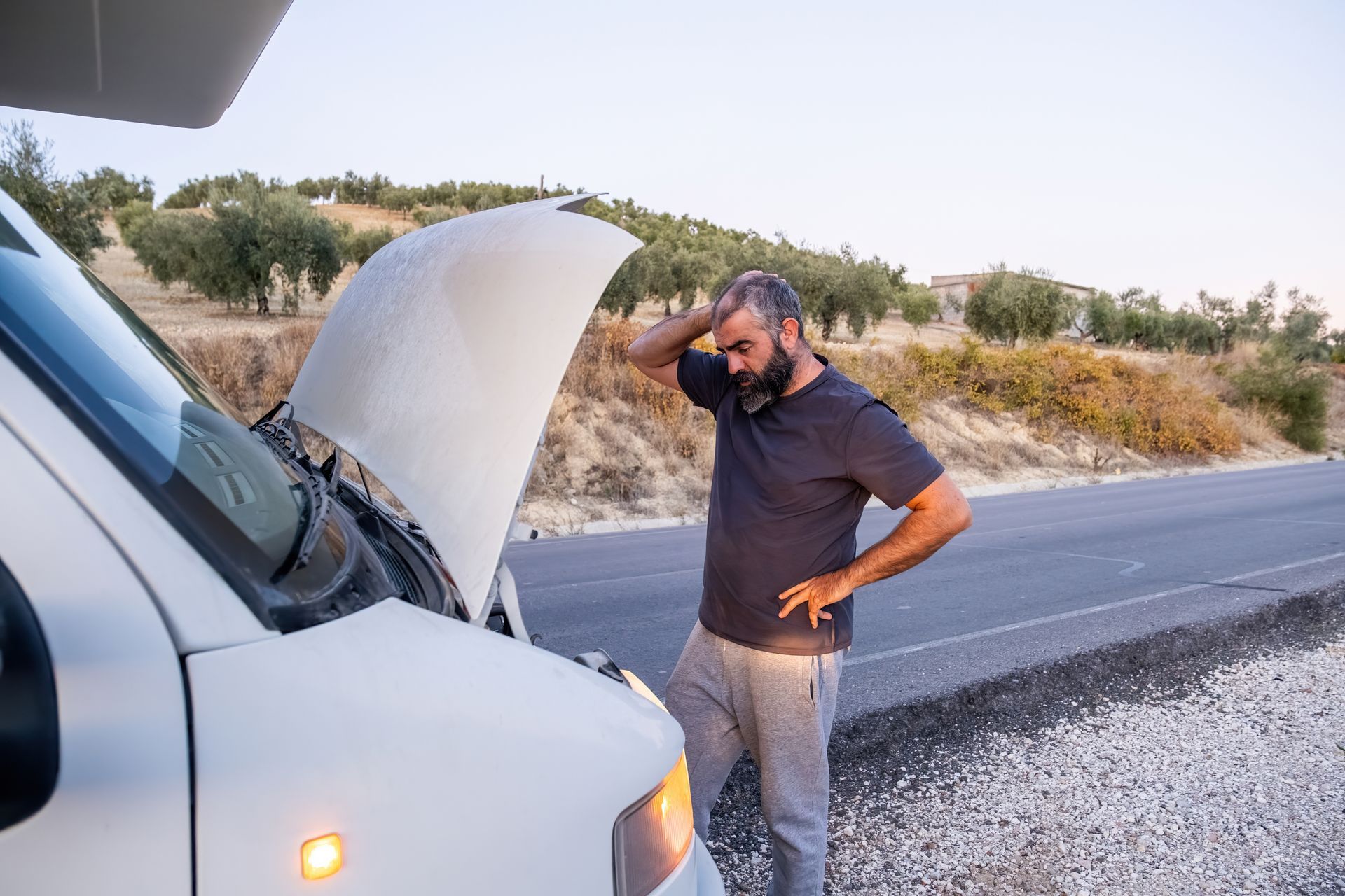 Un homme se tient debout à côté d'une camionnette blanche, capot ouvert, en bord de route, la main sur la tête.