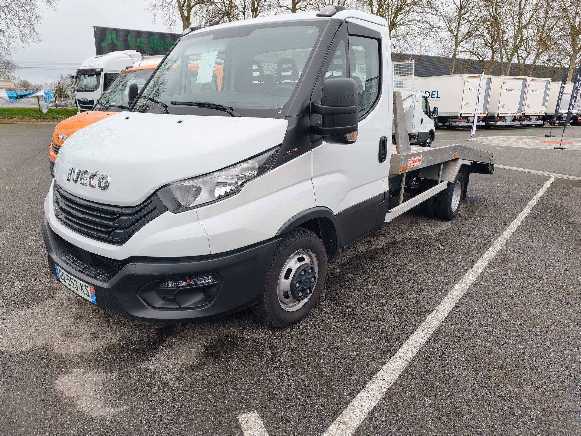 Camion plateau Iveco blanc stationné sur un parking, vue de face en angle.
