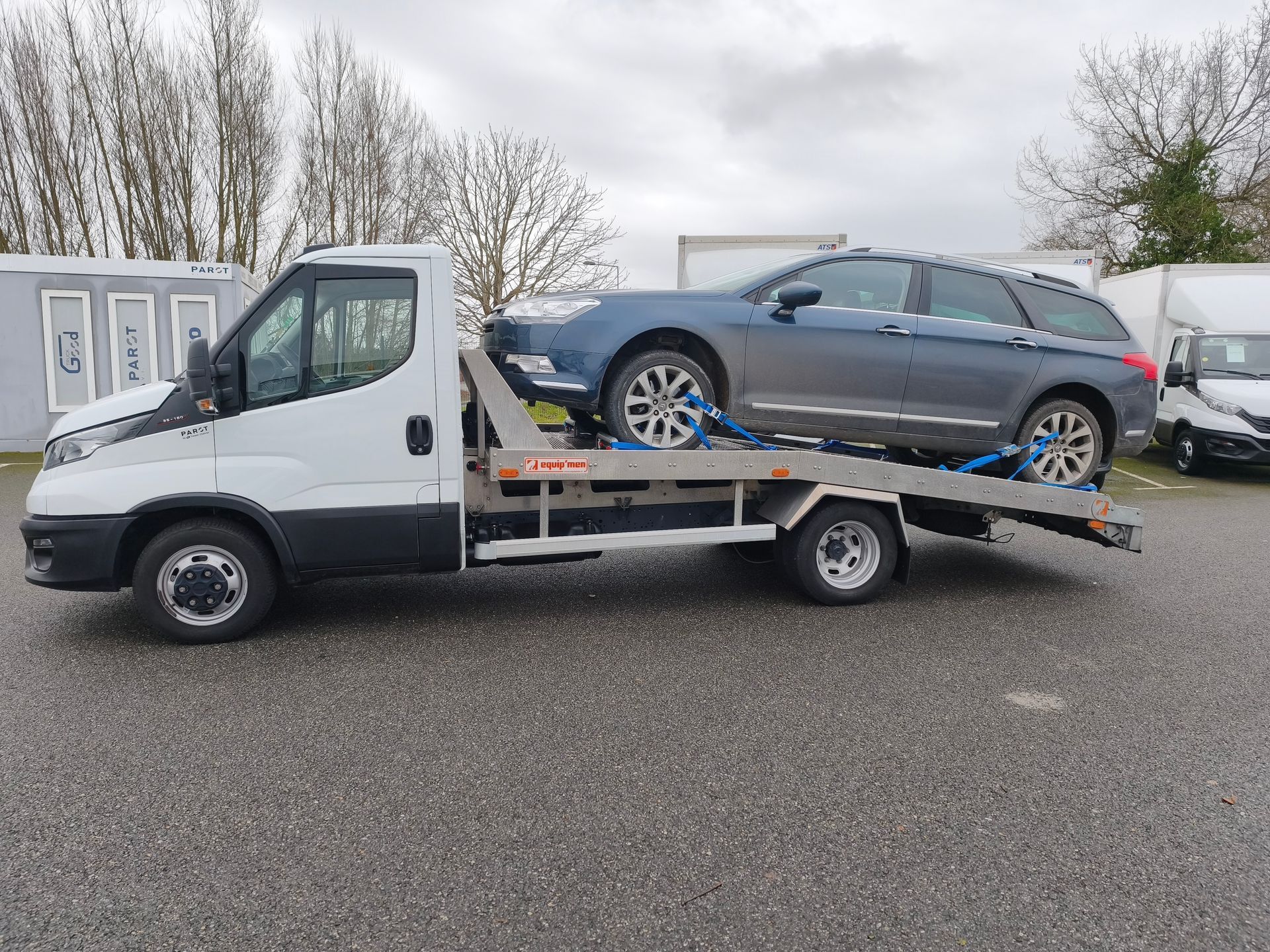 Un camion de dépannage blanc à plateau transportant une berline grise sur un parking extérieur.