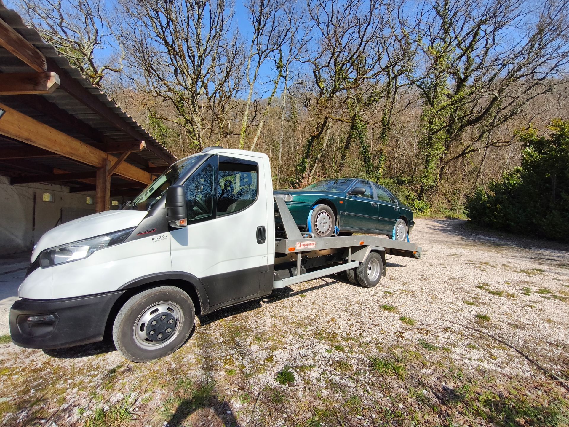 Une dépanneuse blanche transporte une voiture verte sur une allée de gravier, près d'une maison et d'arbres.