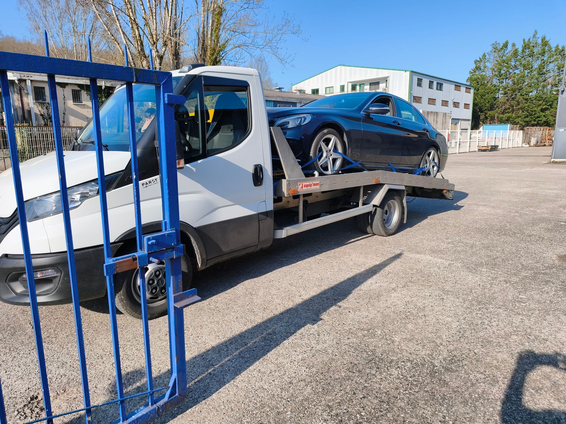 Dépanneuse transportant une voiture sur un parking ensoleillé à côté d'un portail bleu.