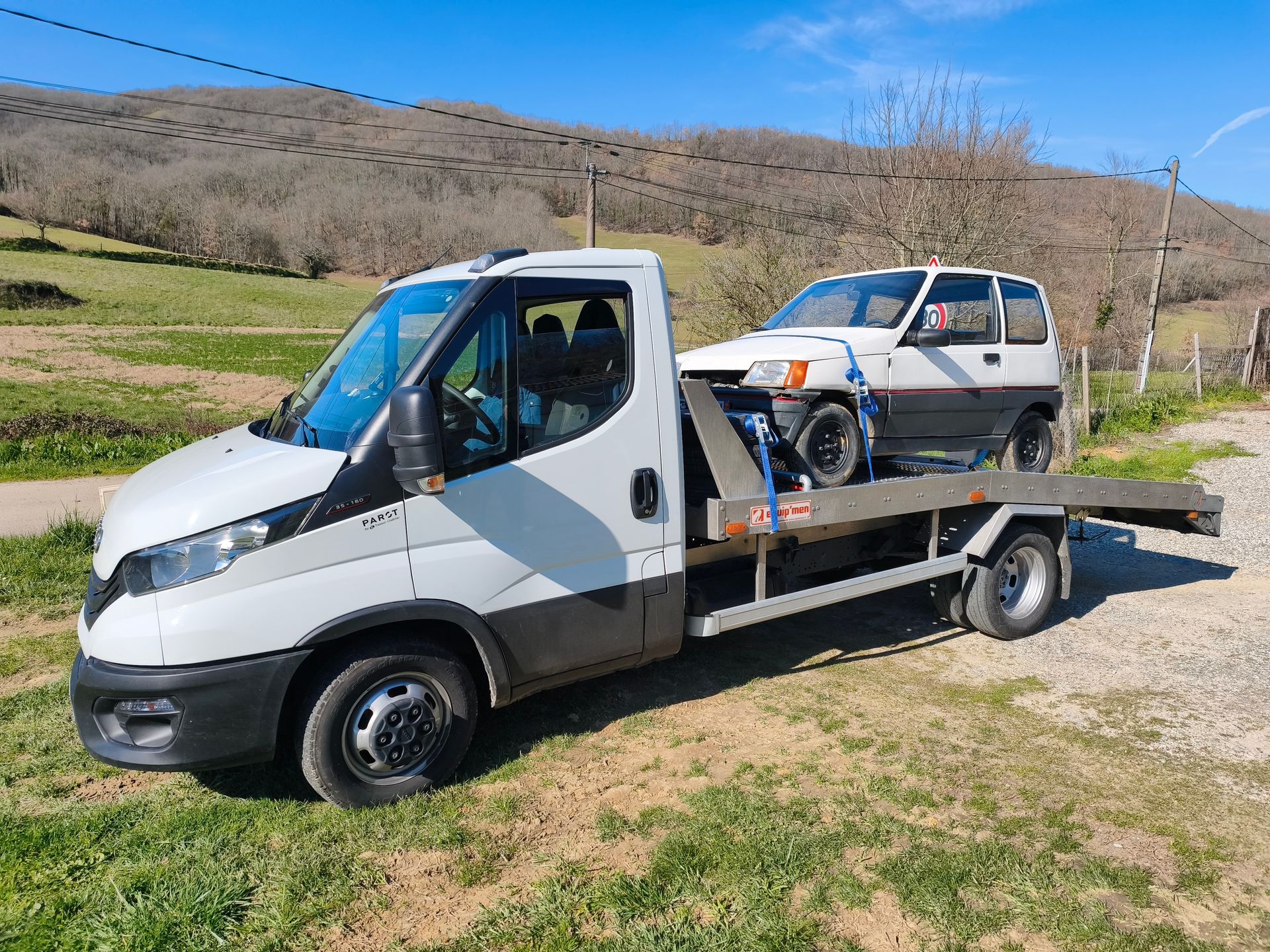 Un camion de dépannage blanc à plateau transporte une camionnette blanche sur le bord d'une route herbeuse, avec des collines en arrière-plan.