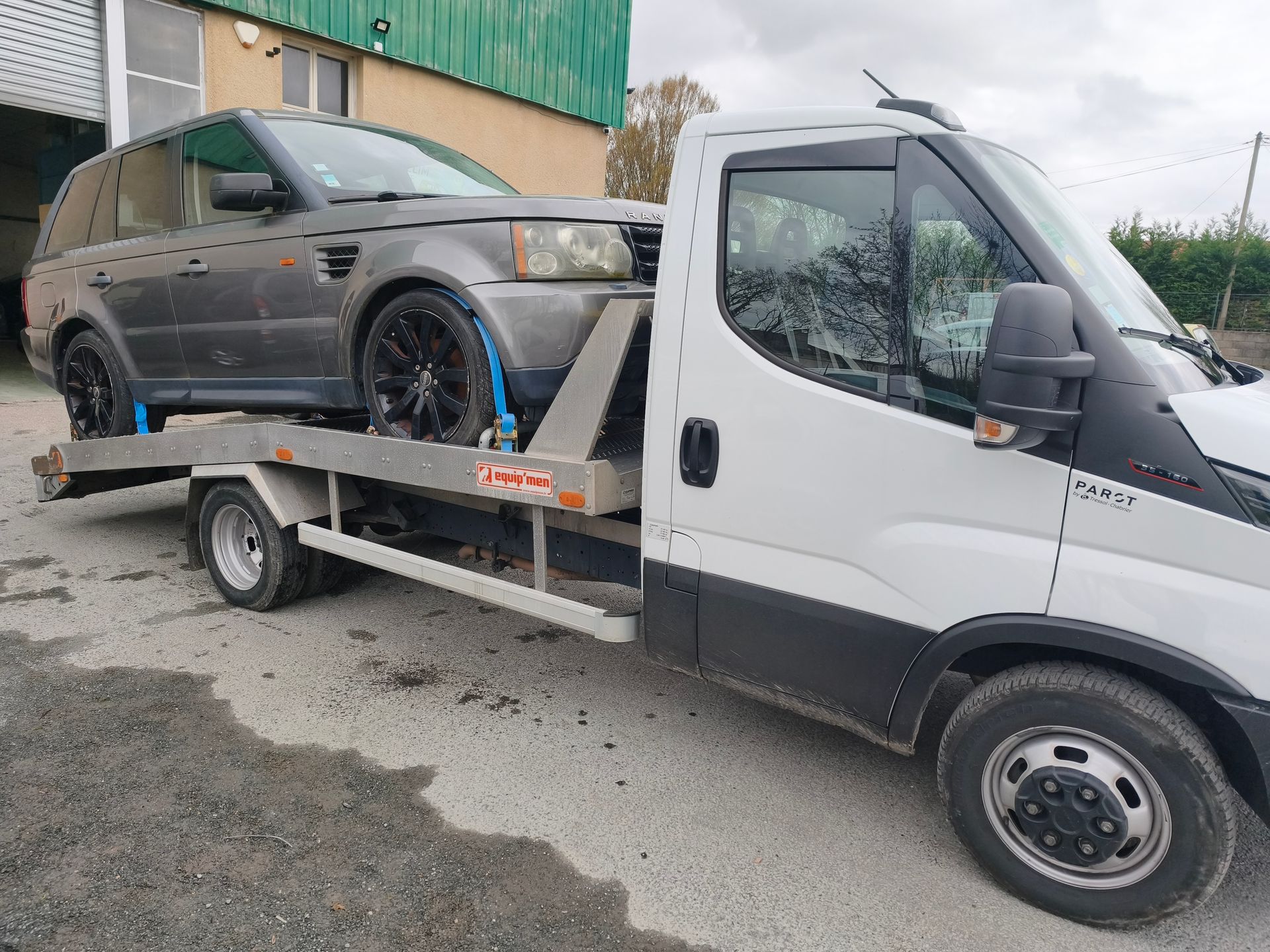 Une dépanneuse blanche transporte un SUV gris sur un plateau à côté d'un bâtiment vert.