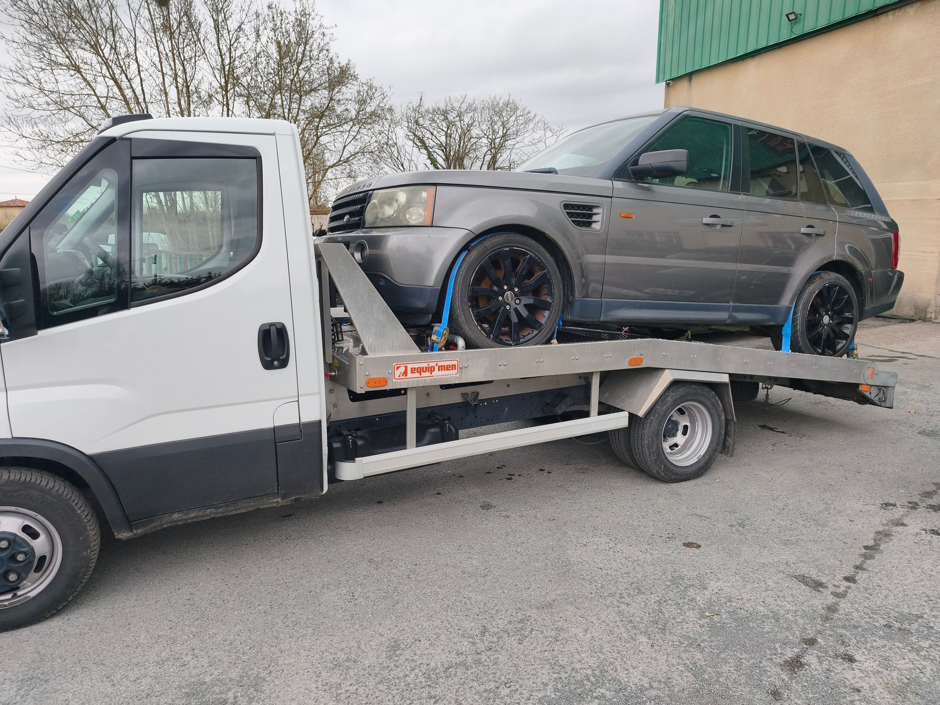 Une dépanneuse transporte un SUV gris sur un plateau à côté d'un bâtiment à l'extérieur.