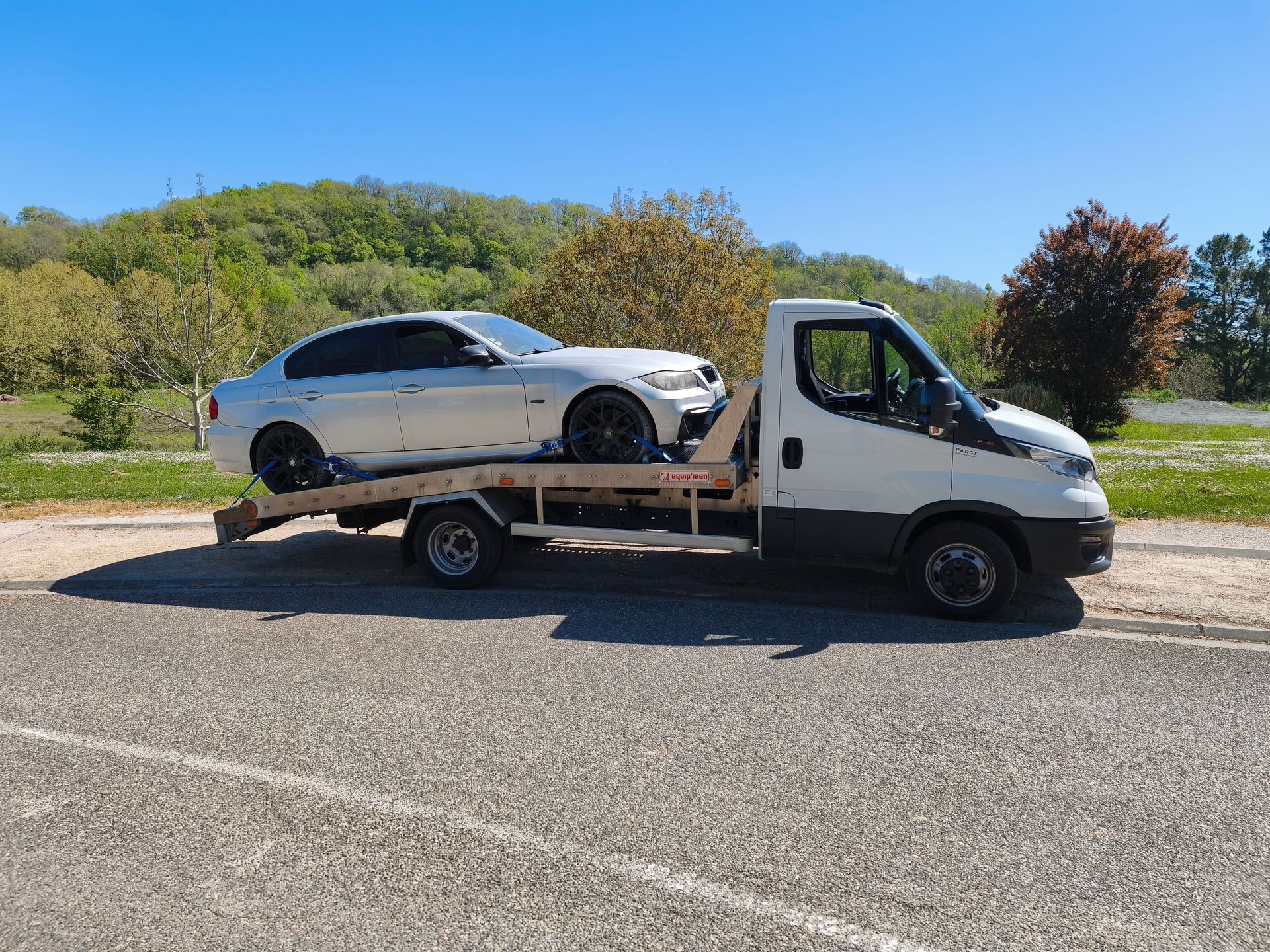 Une voiture chargée sur une dépanneuse à plateau, stationnée en bord de route sous un ciel bleu.