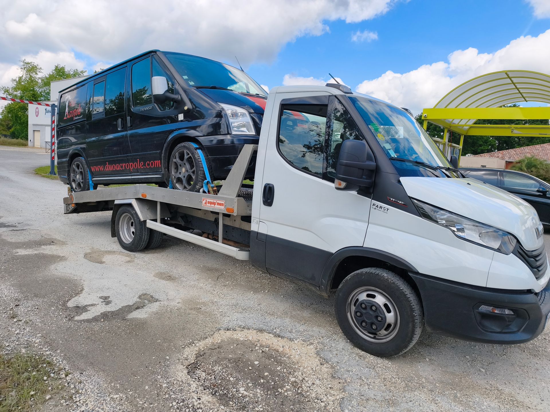 Une dépanneuse transporte un van noir sur un plateau dans un parking sous un ciel nuageux.