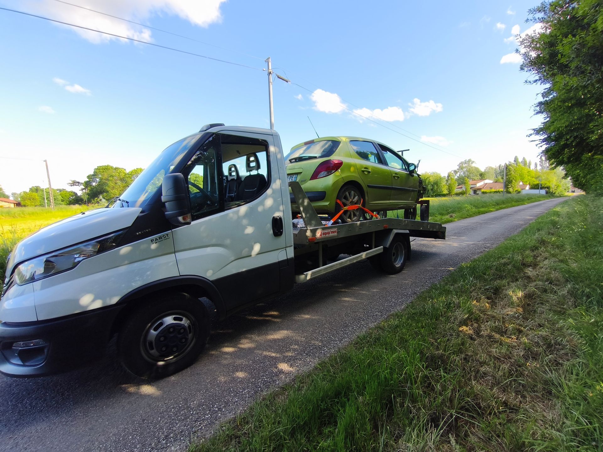 Une dépanneuse transportant une voiture verte au bord d'une route de campagne sous un ciel bleu.