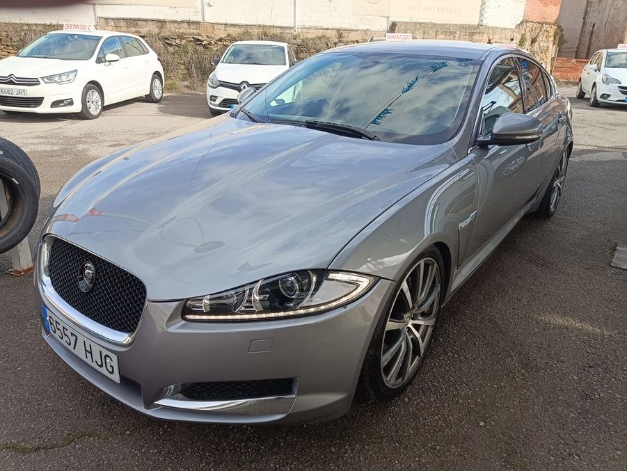 Sedán Jaguar XF plateado estacionado en un lote pavimentado con otros autos al fondo.