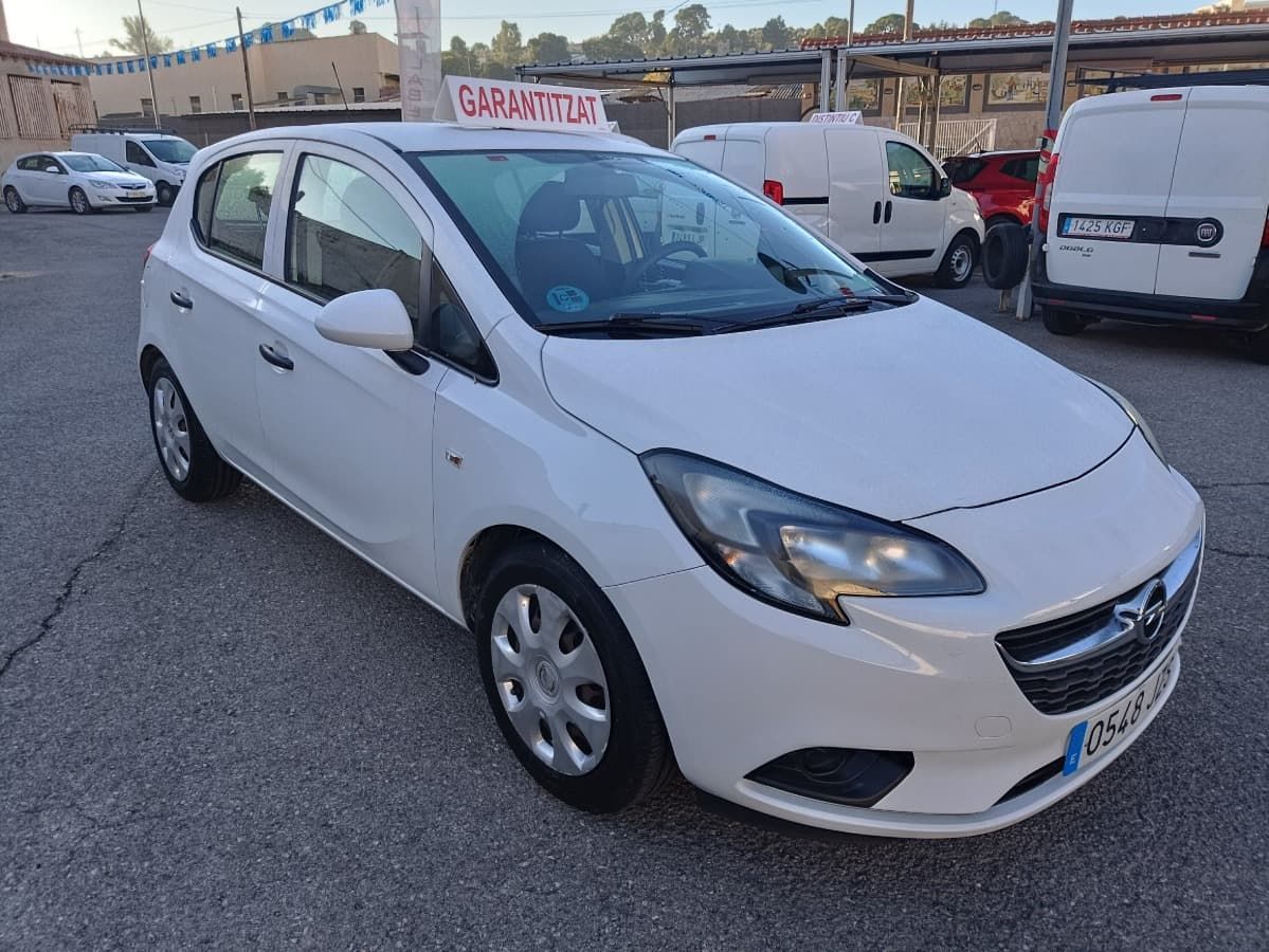 Un Opel Corsa hatchback blanco estacionado en un aparcamiento asfaltado. Al fondo se ven otros coches y furgonetas.