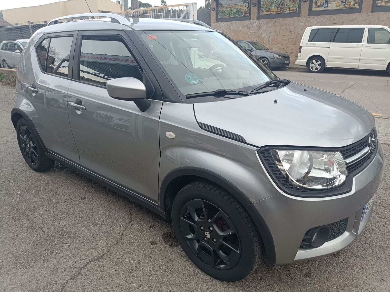 Un Suzuki Ignis gris estacionado en un aparcamiento pavimentado, con llantas de aleación negras y barras en el techo, con un telón de fondo de edificios.